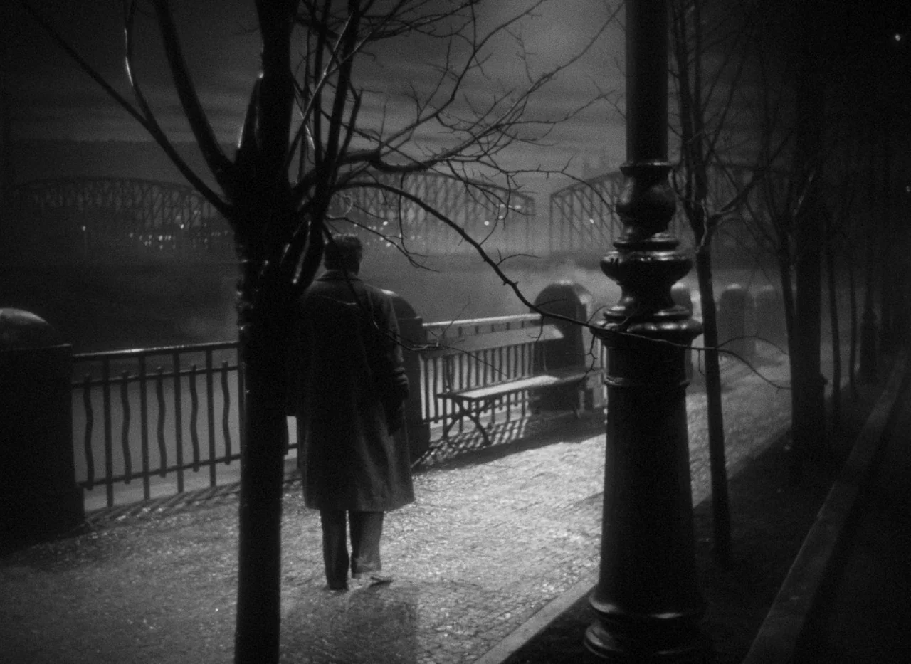 Krakatit (1948) movie still: fullbody — A dark cobblestone embankment beside a river features a silhouette of a man walking. A tall metal lamppost…