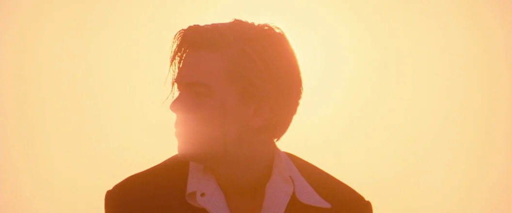 Romeo + Juliet (1996) movie still: closeup — A man stands outdoors silhouetted against a bright, hazy, golden-yellow sky at sunset. The subject is…