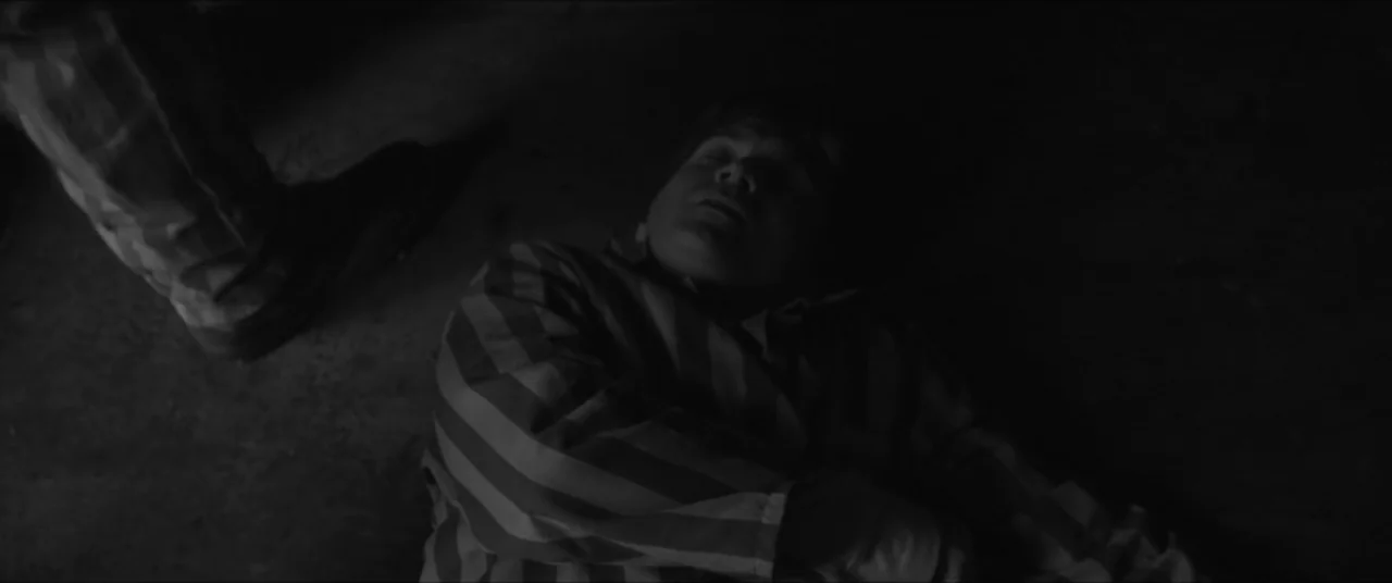 Bau, Artist at War (2025) movie still: medium — A dimly lit room features a person on the floor wearing striped clothing. A large boot is positioned…