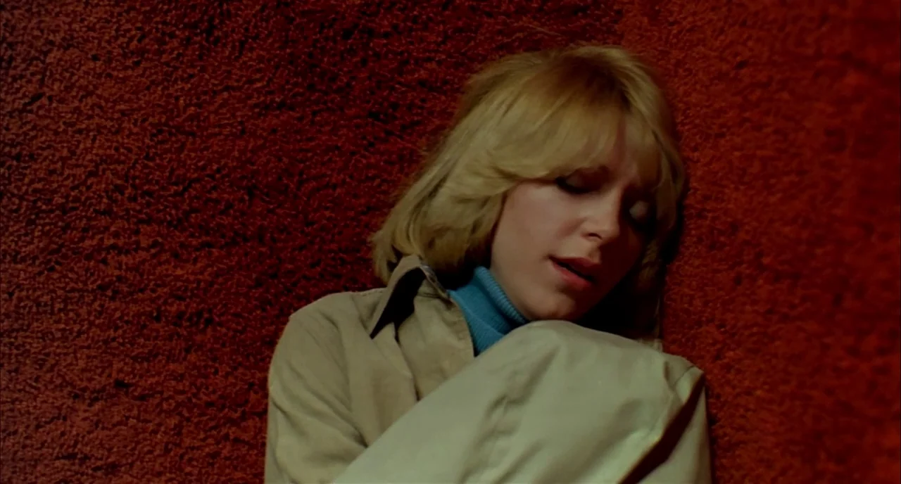 Dawn of the Dead (1978) movie still: closeup — A woman leans against a vibrant, deep red carpeted wall in a corner. She sits with her knees pulled…
