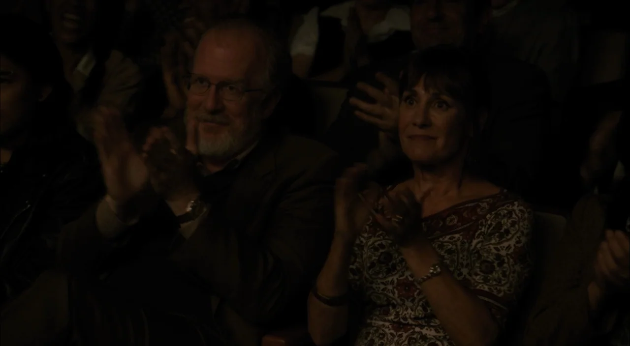 Lady Bird (2017) movie still: medium — A dimly lit auditorium filled with people seated in rows. An older man and a woman in the foreground clap…