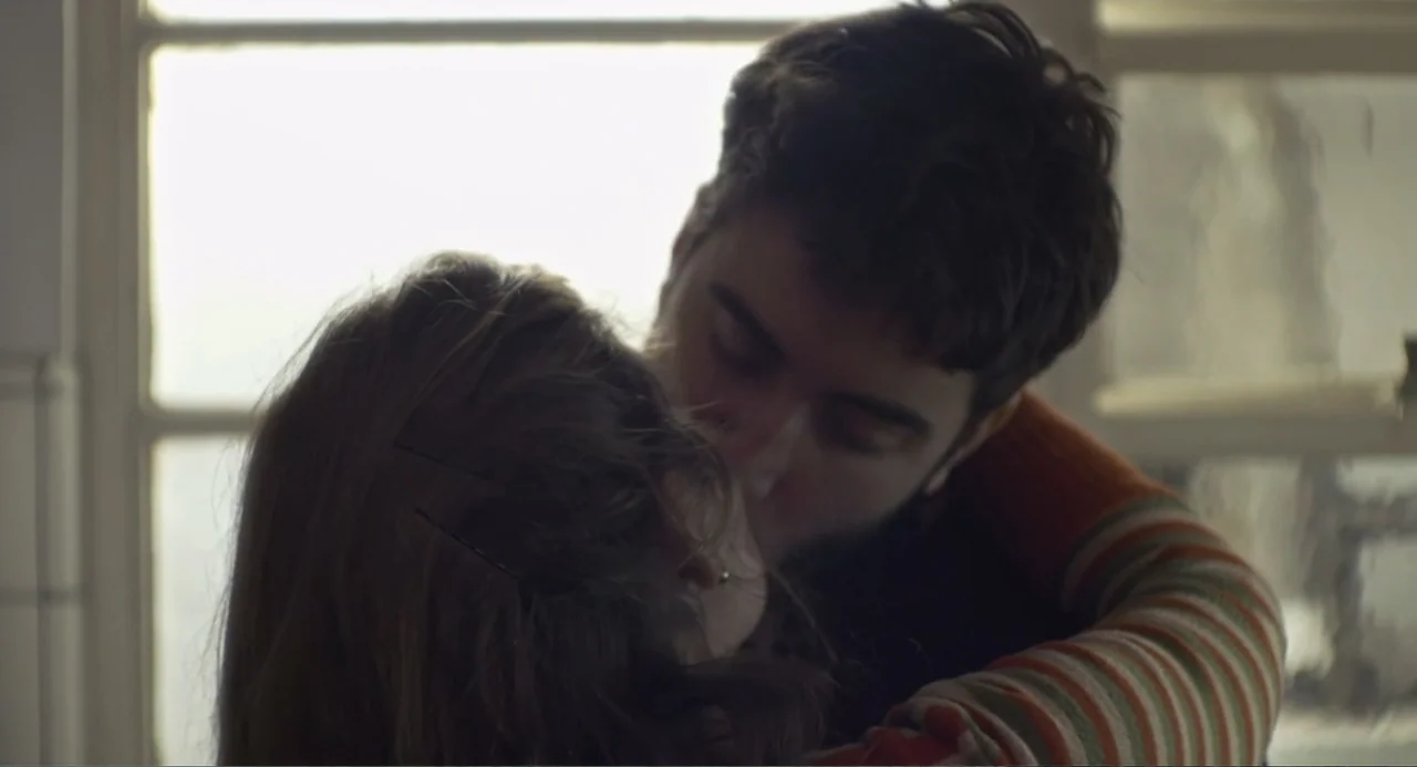 An Odd Turn (2024) movie still: closeup — An interior space features a bright window frame in the background. A young couple embraces and kisses in…