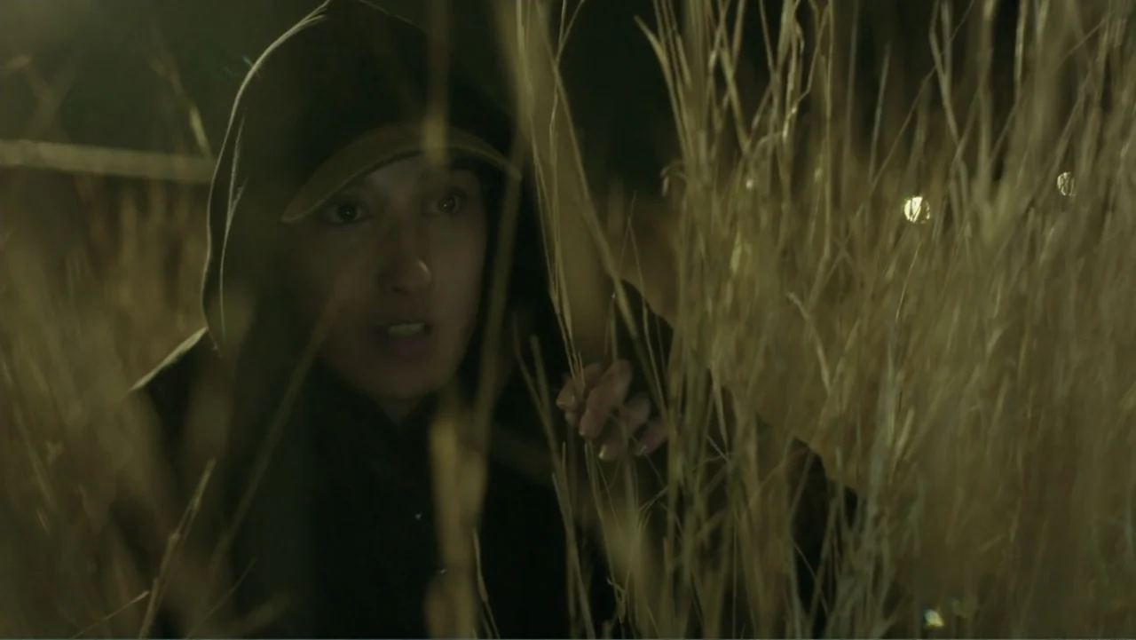 Code of Trust (2019) movie still: closeup — A dark night setting featuring dense, tall stalks of dry grass in an open area. A young woman with a…