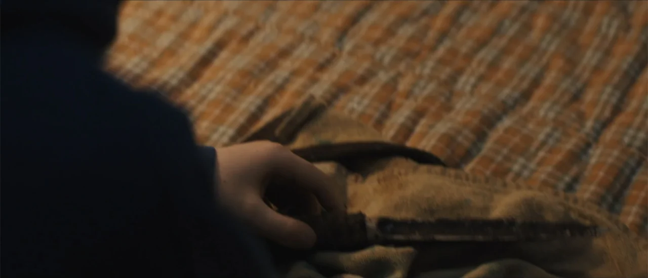 All Fun and Games (2023) movie still: closeup — A plaid fabric blanket rests on a soft surface in a dim room. A hand reaches into the frame to touch…