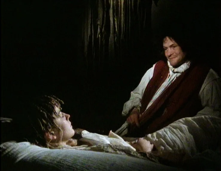 The Tempest (1979) movie still: medium — A dark, spartan bedroom contains a bed with heavy white linens. A man sits beside a woman lying…