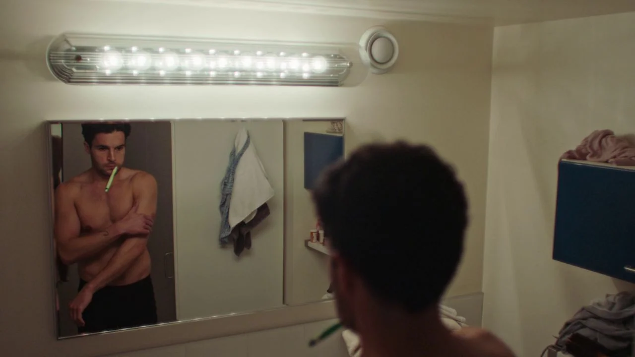 Possessor (2020) movie still: medium — A man observes himself in a wall mirror inside a brightly lit tiled room. The man stands shirtless with a…