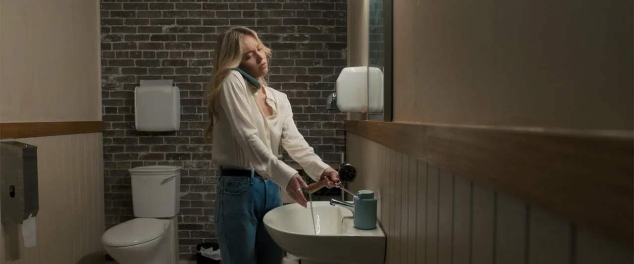 Anyone But You (2023) movie still: medium — A tiled restroom features a dark brick wall and a white porcelain sink. A young woman stands at the sink…