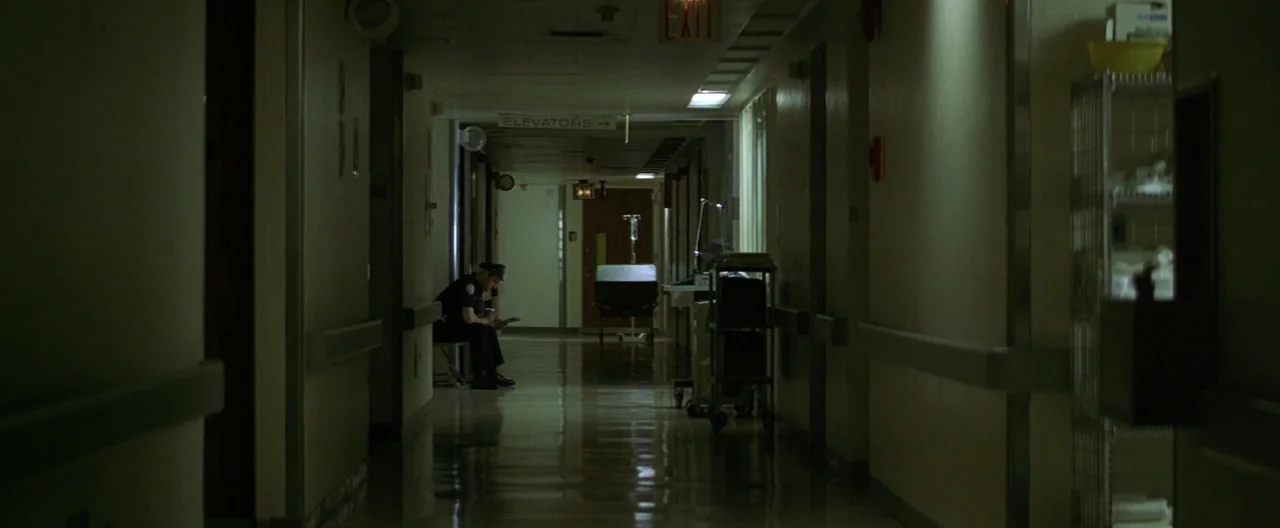 The Yards (2000) movie still: wide — A long dimly lit medical hallway features linoleum flooring and cream-colored walls. A lone police officer sits…