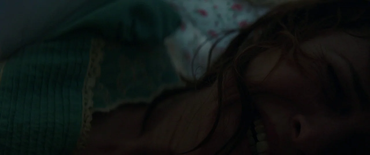 Pet Sematary (2019) movie still: closeup — A close-up of a woman lying in a dimly lit room with floral bedding. She is positioned with her teeth…