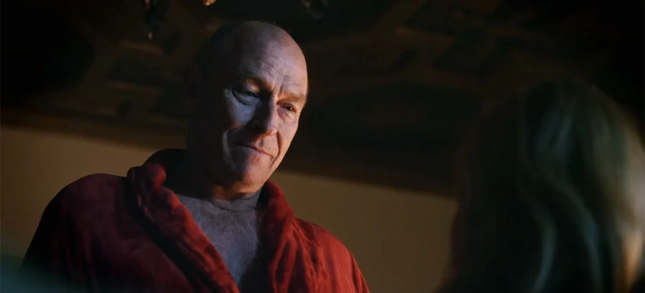 The Russian Bride (2019) movie still: medium — A dimly lit indoor space with low-key illumination highlights a bald older man. The man wears a red…