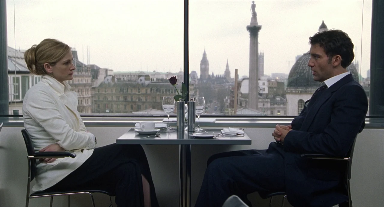 Closer (2004) movie still: wide — A modern restaurant interior with a large window looking out over a city landscape. A man and a woman sit facing…