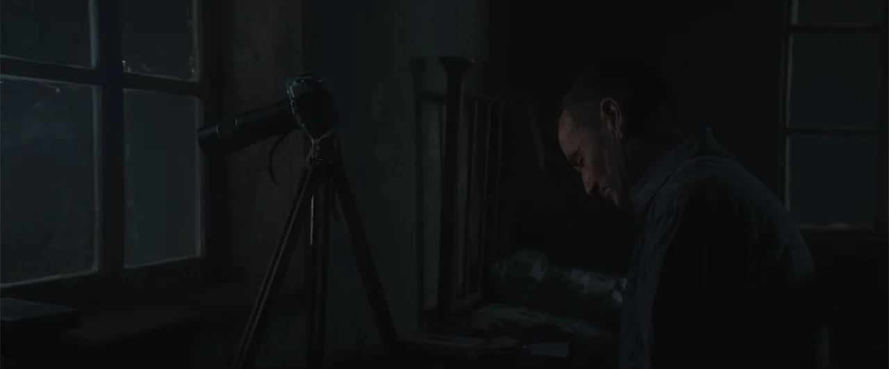 My Neighbor Adolf (2022) movie still: medium — A dark, spartan room contains a camera on a wooden tripod near a window. An elderly man sits in the…