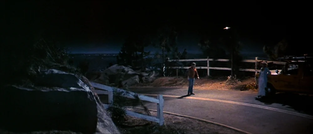 Close Encounters Of The Third Kind (1977) movie still: wide — A winding mountain road at night with rocky embankments and a wooden fence. A man…
