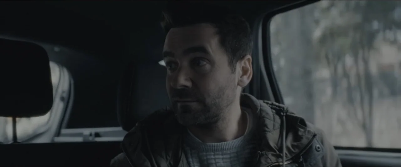 Quicksand (2023) movie still: medium — A man sits in the back seat of a vehicle during the day. He looks toward the side window while wearing a…