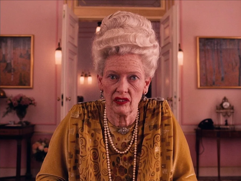 The Grand Budapest Hotel (2014) movie still: medium — A formal room features pink walls, symmetrically hung paintings, and double doors in back. An…