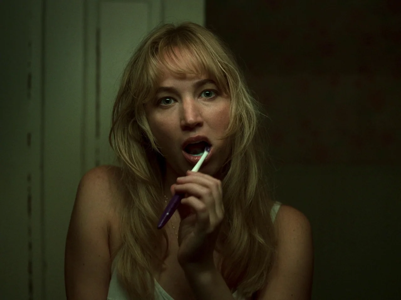 Die My Love (2025) movie still: closeup — A dark, dimly lit bathroom space with muted, greenish tones. A woman brushes her teeth while looking…