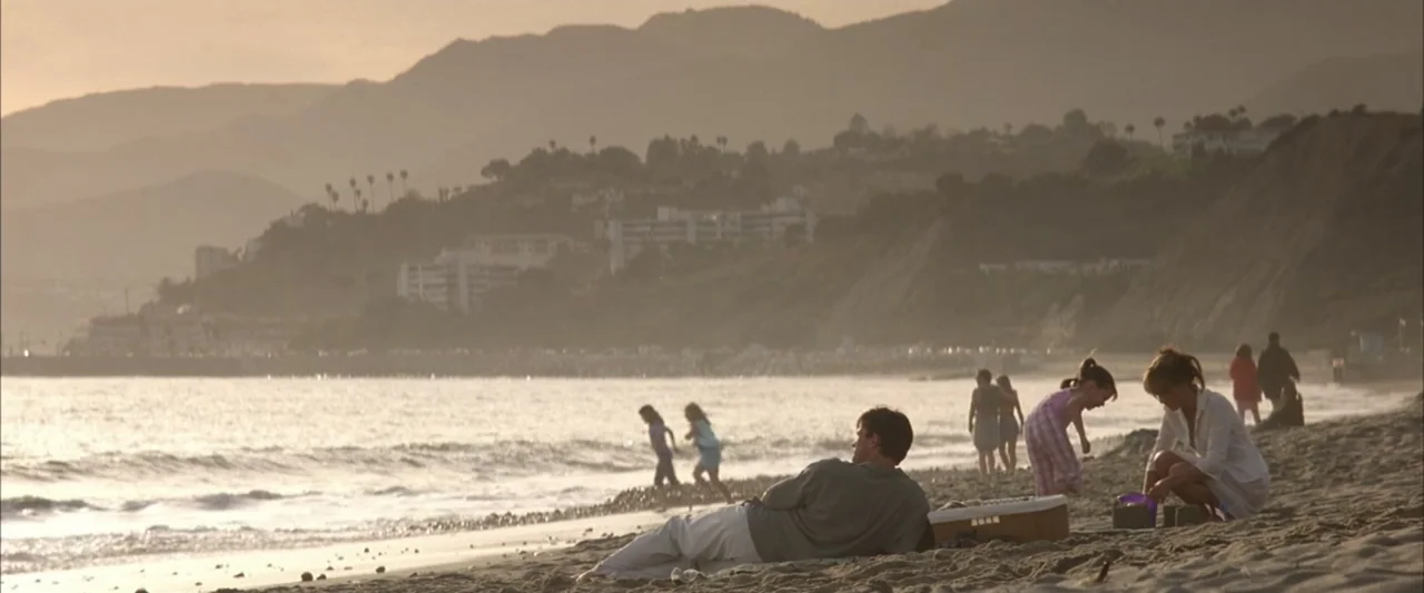Enough (2002) movie still: wide — A wide beach features sand in the foreground and hilly coastal development behind it. A group of people lounge and…