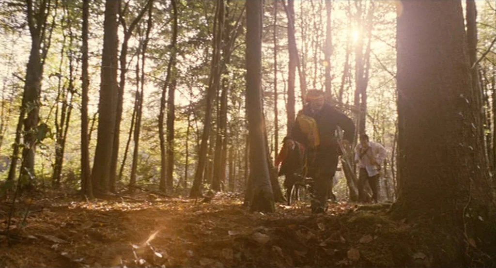 Submarine movie still: wide — A dense forest with tall trees and leaf-covered ground is illuminated by bright sunlight. Three individuals walk…