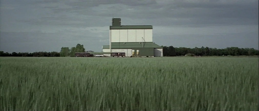 Time Of The Wolf (2003) movie still: establishing — A tall white and green grain elevator stands isolated in the middle of a field. Green wheat…