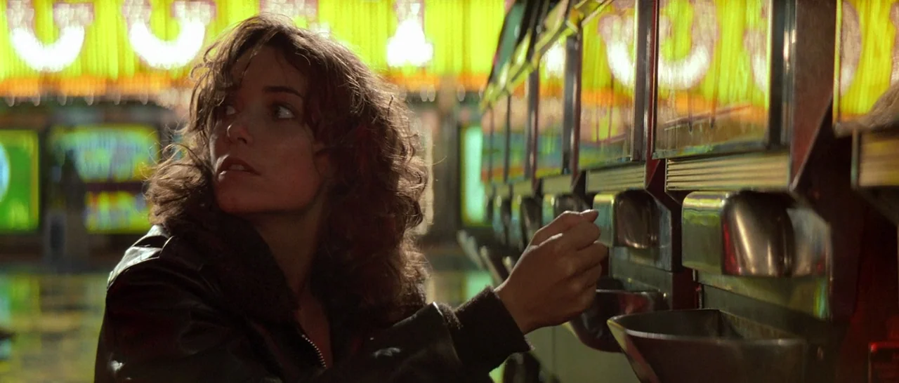 Starman (1984) movie still: medium — An arcade interior glows with repetitive yellow neon lights along a wall of machines. A woman with wavy brown…