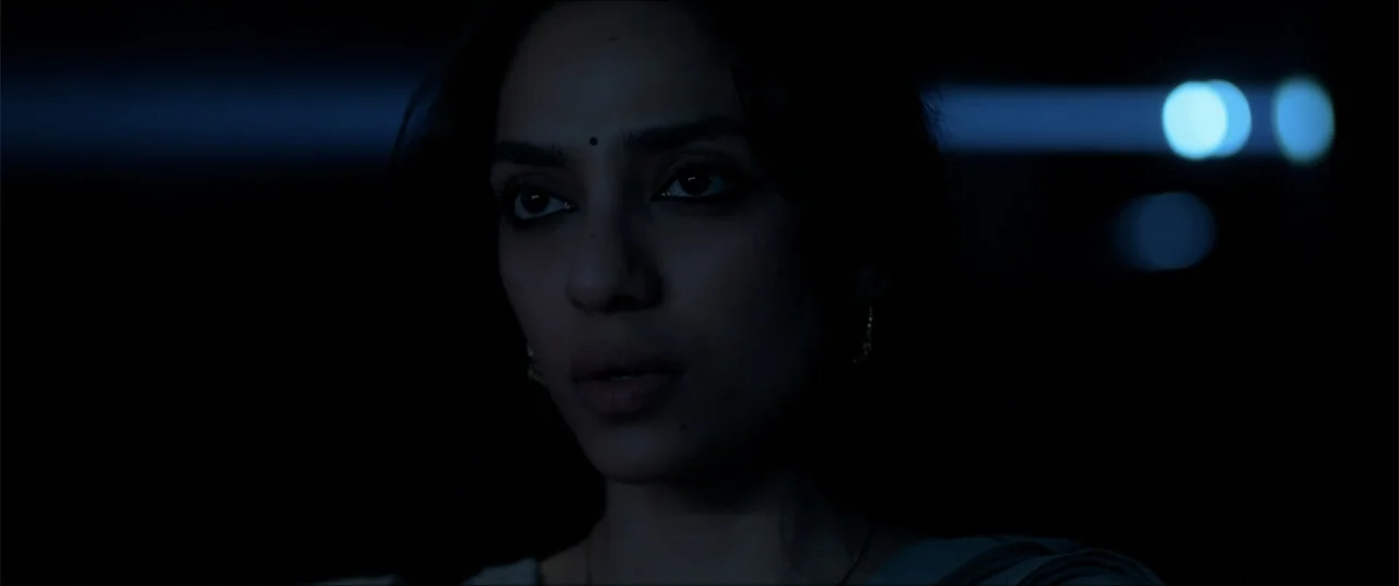Cheekatilo (2026) movie still: closeup — A dark vehicle interior with a woman centered in the frame. The woman looks forward while the blue light…