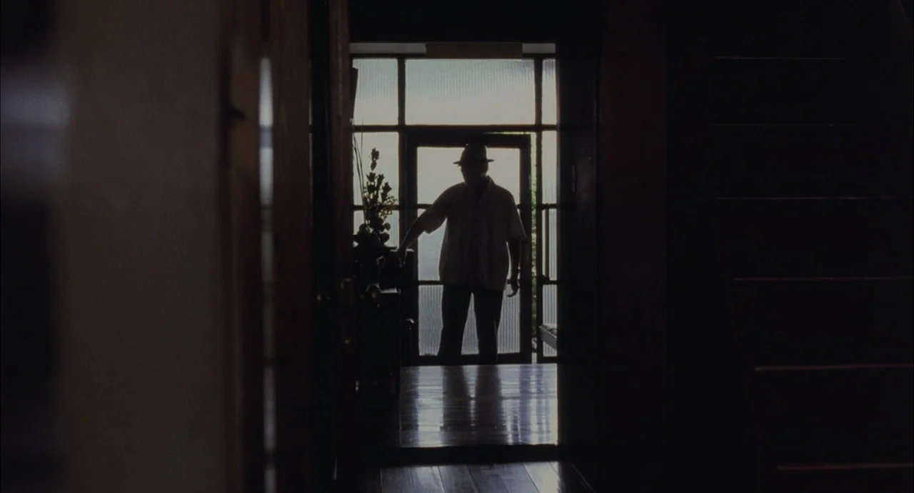 Still Walking (2008) movie still: medium — A dark hallway leads toward a bright doorway framed by glass panes. A silhouette of a man in a hat stands…