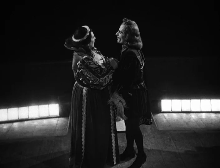 The White Sheik (1952) movie still: fullbody — Two figures in Renaissance-style period clothing stand on a dark, minimalist stage platform. Bright…