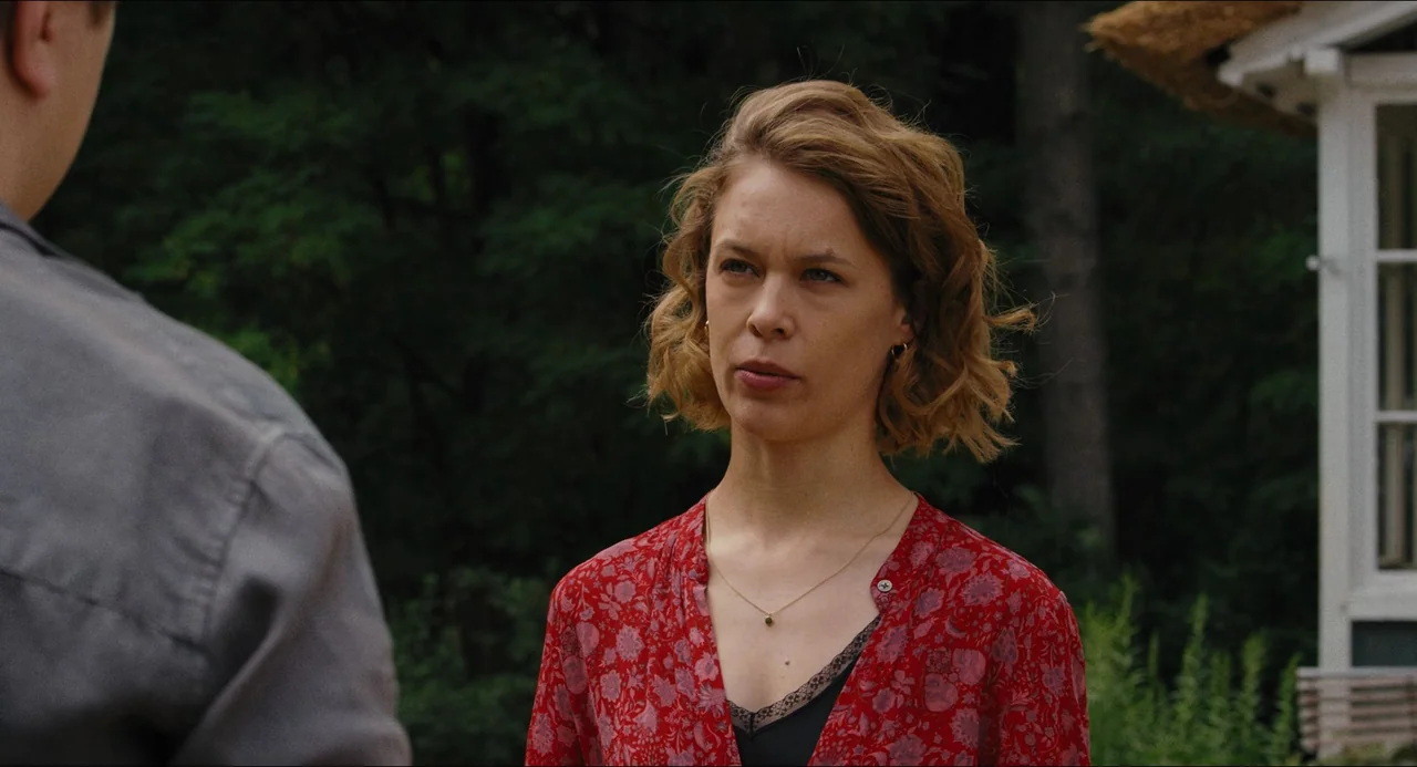 Afire (2023) movie still: medium — An outdoor area with a blurred green tree line and a white building corner. A woman with wavy blonde hair stands…
