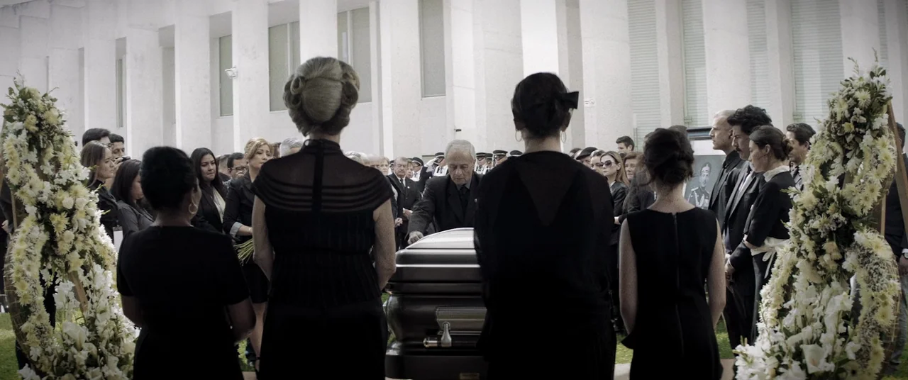 La Llorona (2019) movie still: wide — A group of mourners in dark attire stands in an open-air plaza with pillars. They surround a metallic coffin…