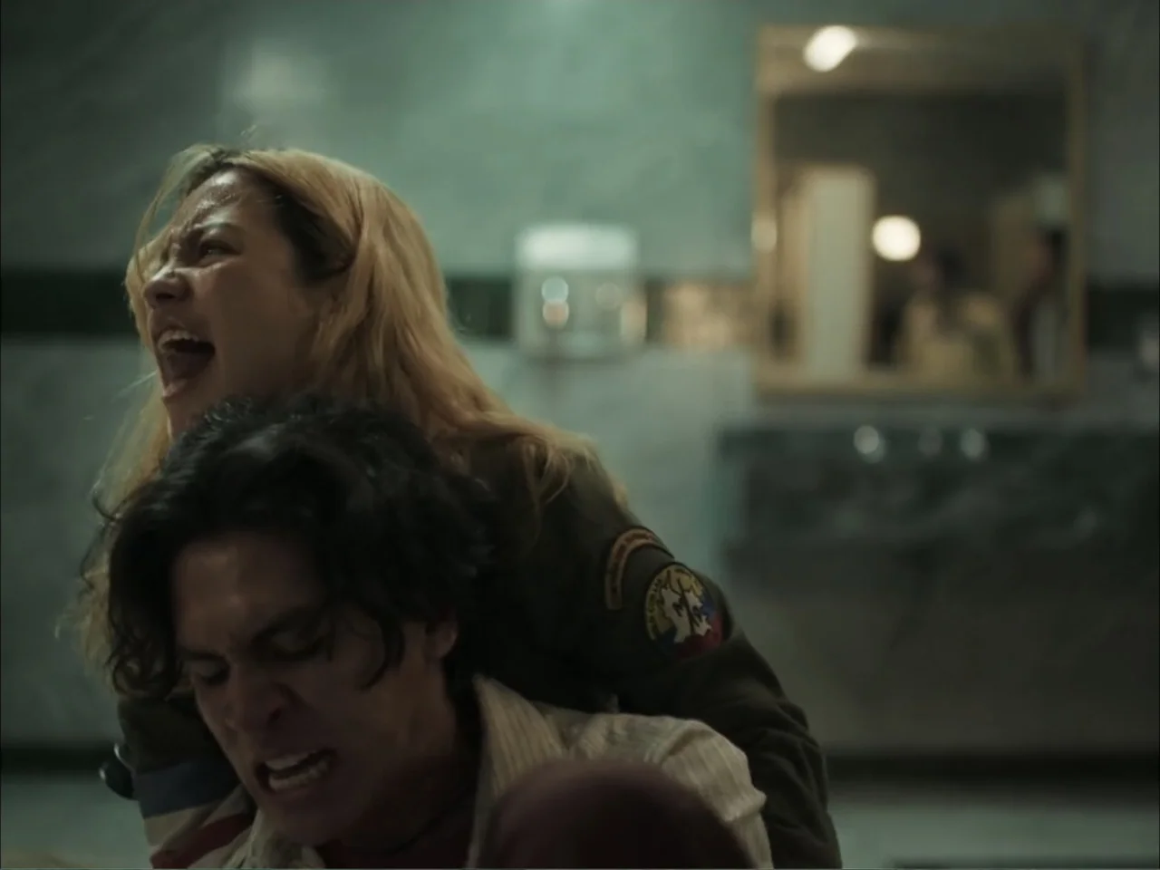 November (2025) movie still: medium — A dimly lit restroom with marble walls and a central vanity mirror visible. A woman screams while holding a…