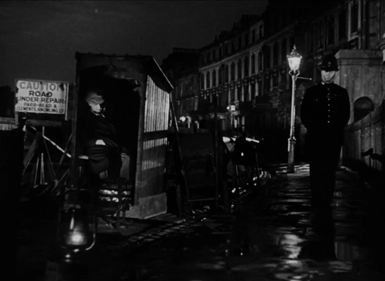The Blue Lamp (1950) movie still: wide — A wet night street in London features a Victorian-style gas lamp and row houses. A policeman walks past a…