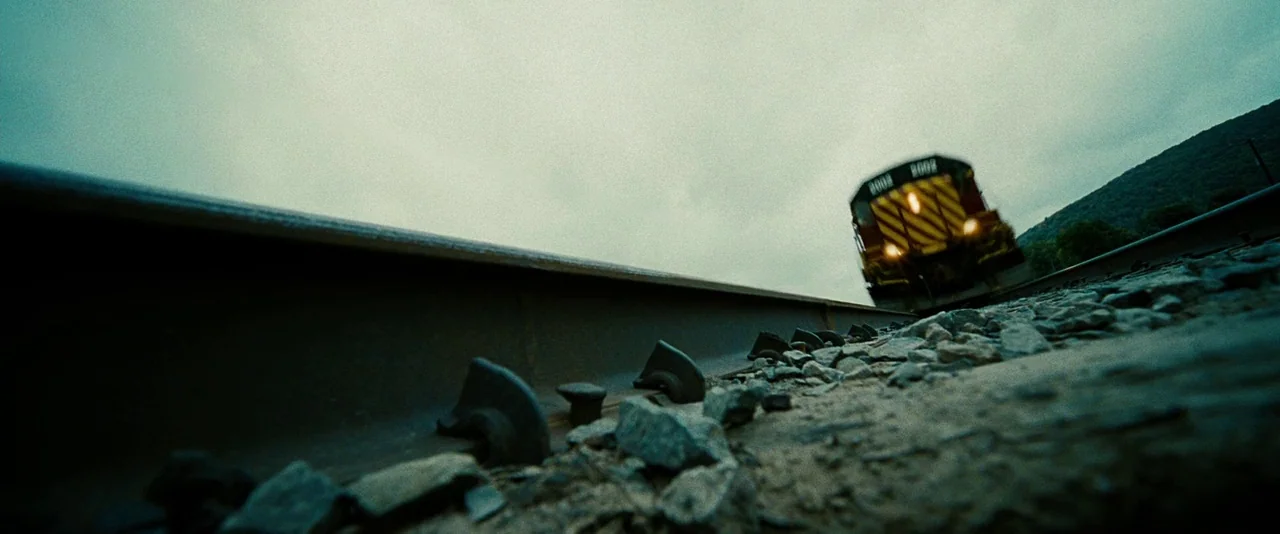 Unstoppable (2010) movie still: wide — A low-angle view rests on gravel train tracks under a cloudy sky. A diesel locomotive approaches in the…
