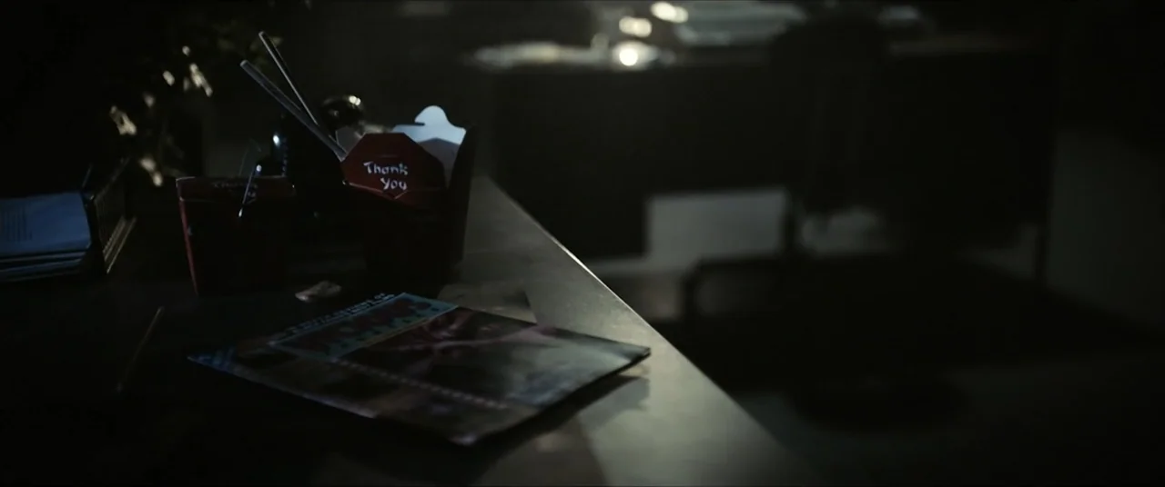 Vicious Fun (2021) movie still: closeup — A cluttered dark desk surface holds a takeout container and a magazine. The rest of the workspace remains…