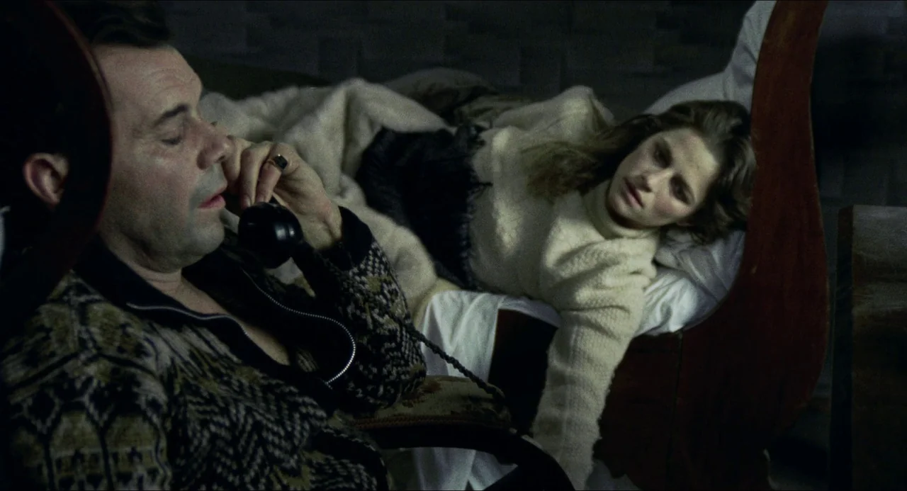 The Night Porter (1974) movie still: medium — A dimly lit room containing a man in a chair and a woman on a bed. The man speaks on a black telephone…