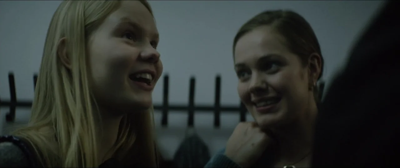 Dogs Don't Wear Pants (2019) movie still: medium — A dimly lit room features a dark metal coat rack mounted on a wall. Two young women stand near…