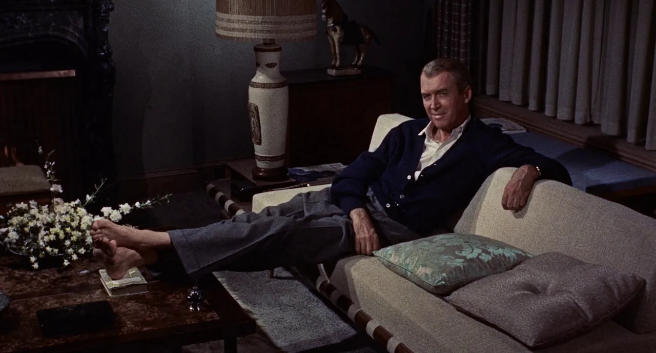 Bell Book and Candle (1958) movie still: medium — A dimly lit room with mid-century modern furniture and dark wallpaper. A man lies on a beige couch…