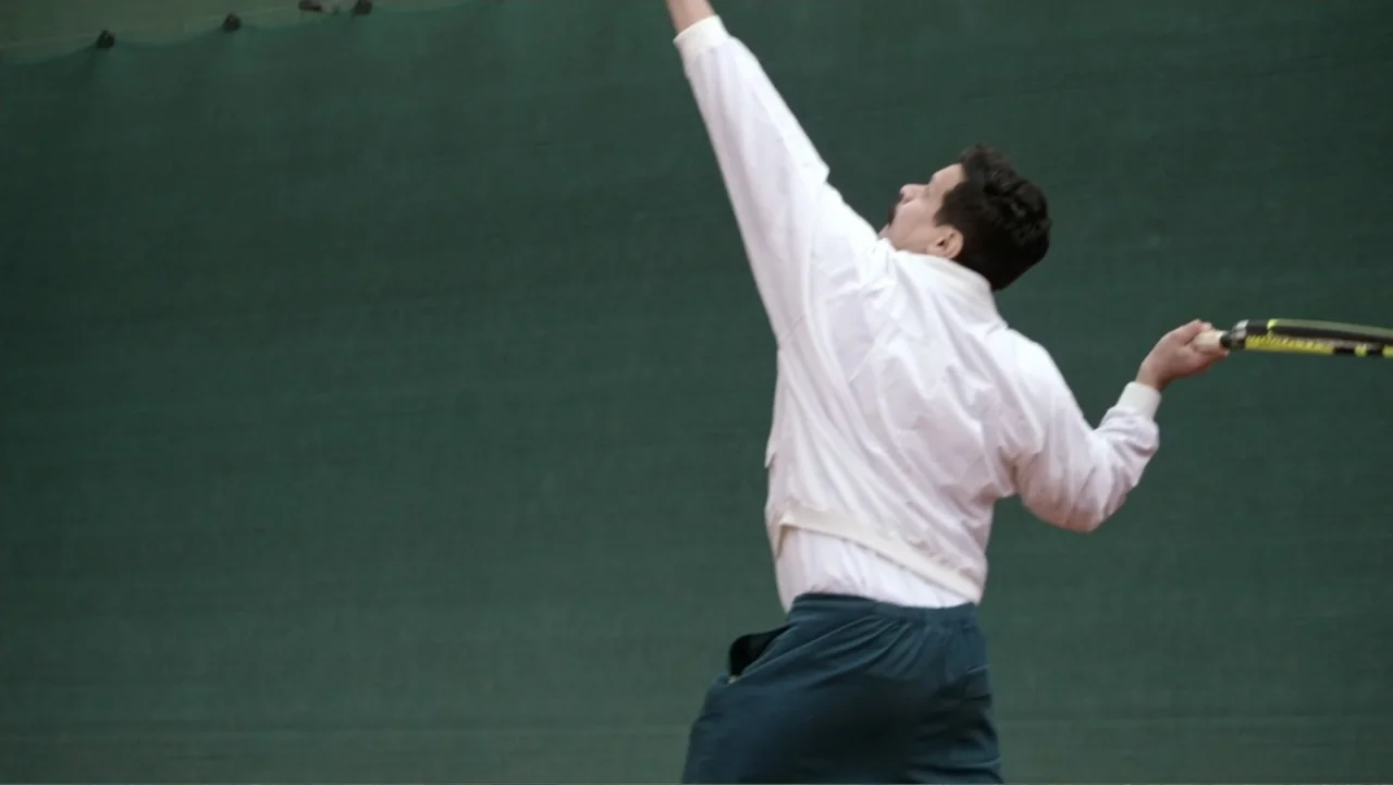 Saturdays Disorders (2022) movie still: medium — A man stands on a tennis court with a large green privacy screen behind him. The man is mid-serve…