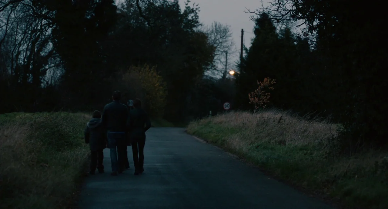 Everyday (2012) movie still: wide — A quiet rural road is bordered by dense trees and tall dried grasses. Three people walk away from the viewer…