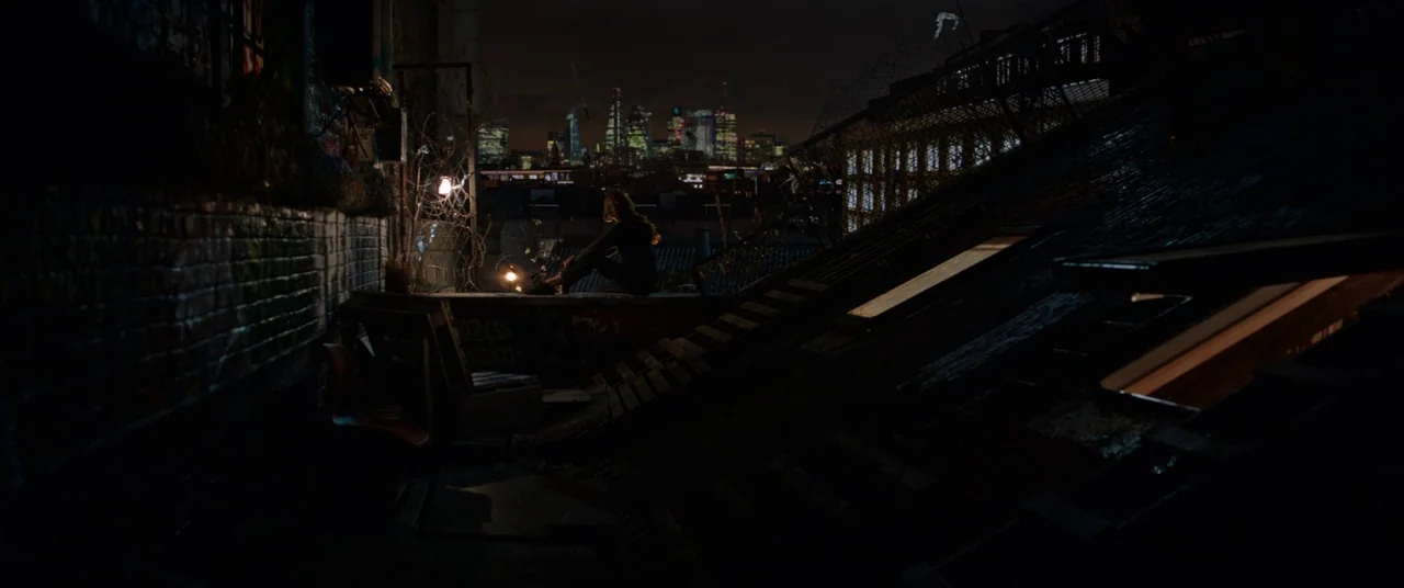 Tomb Raider (2018) movie still: wide — A dark urban rooftop features a brick wall covered in graffiti and wire fencing. A lone figure sits on the…