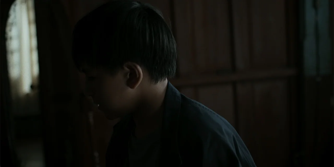 Nokturno (2024) movie still: closeup — A dimly lit room with dark wooden walls provides a textured background. A young boy stands facing the side…