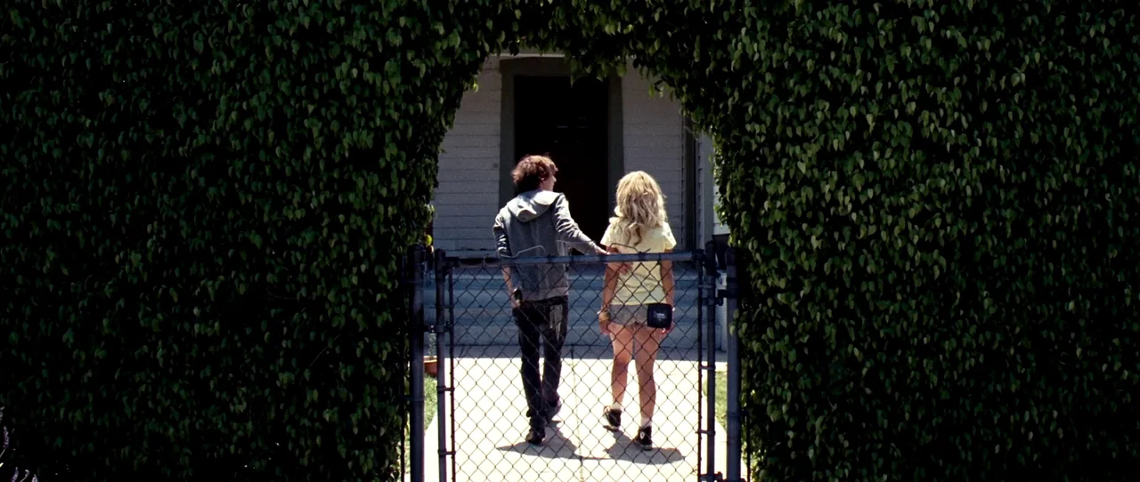 Little Birds (2011) movie still: fullbody — A residential front yard is framed by a dense, manicured archway of green leaves. A young couple walks…
