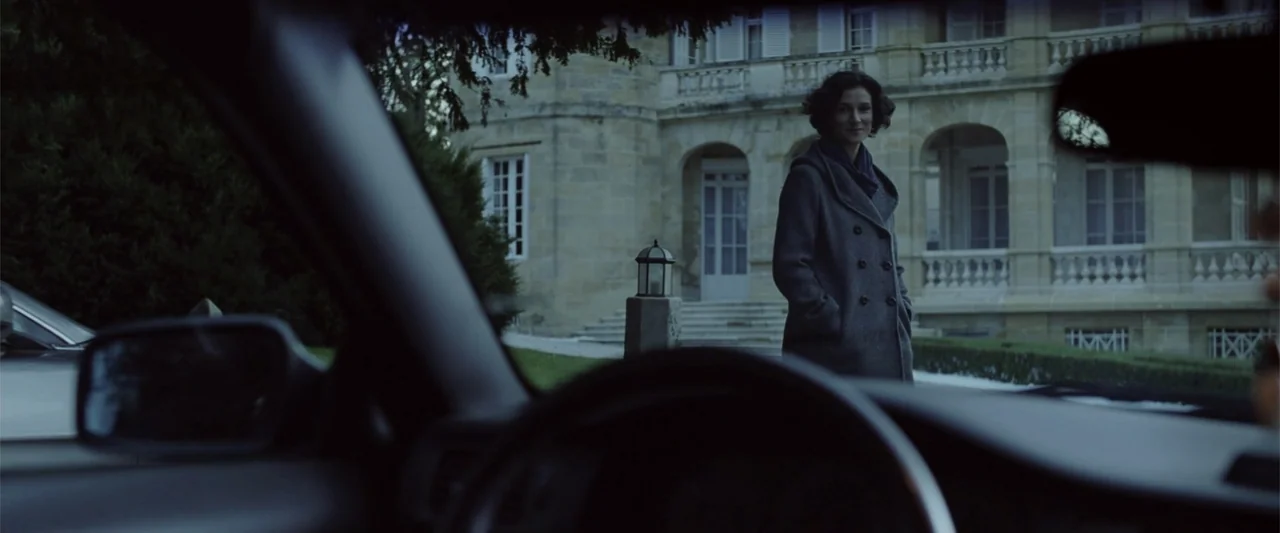 Anna (2013) movie still: medium — A woman stands in the driveway of a large, pale stone mansion at dusk. She is positioned outside a vehicle,…