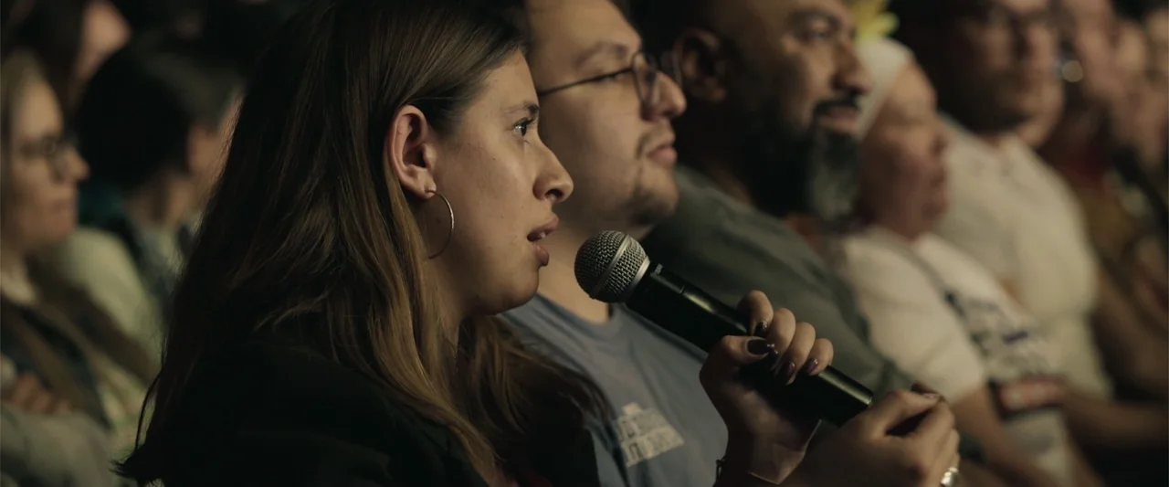 Making of: F*CKS NEWS (2025) movie still: medium — A dimly lit room with rows of people seated for a public presentation. A woman in the foreground…