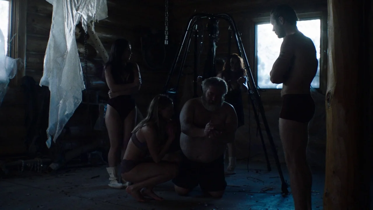 Snowbound (2017) movie still: wide — A dimly lit wooden cabin interior with exposed log walls and scattered debris. A bearded man kneels on the…