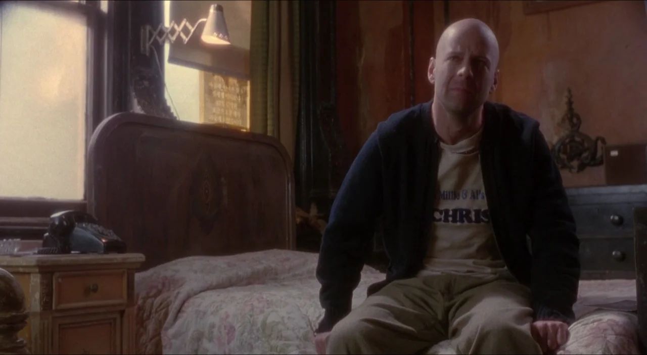 Twelve Monkeys (1995) movie still: medium — A sparsely furnished room features wood paneling and aged wallpaper behind a simple bed. A bald man…