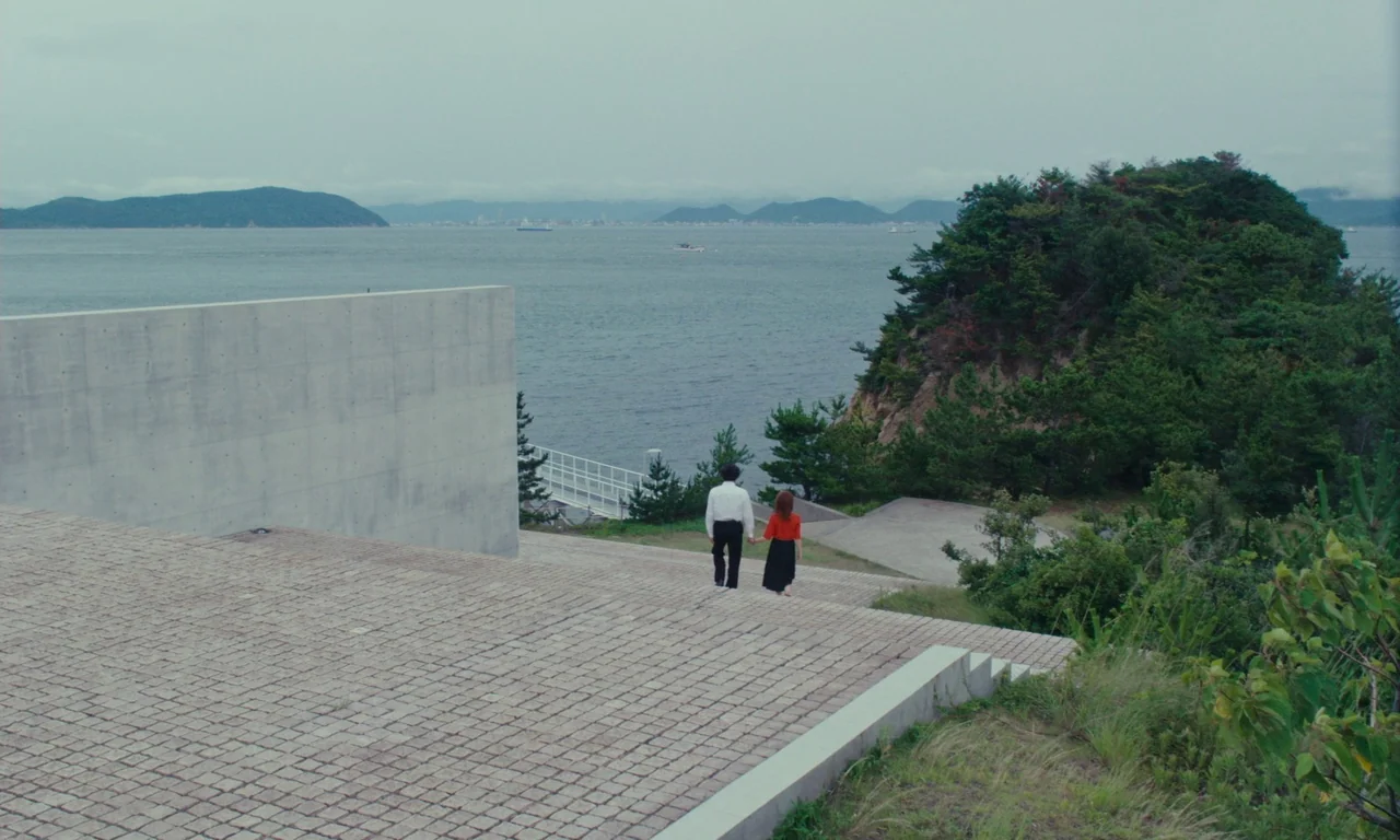 Sidonie in Japan (2023) movie still: wide — An open stone terrace overlooks a vast bay surrounded by distant hills and trees. A couple walks…