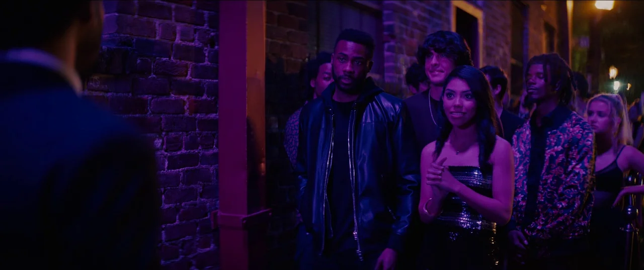 Night Teeth (2021) movie still: medium — A group of people waits in a line against a weathered red brick exterior wall. The atmosphere is heavily…