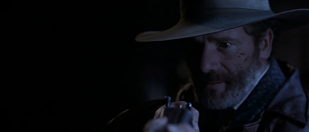 The Claim (2000) movie still: closeup — A man in a wide-brimmed hat stands in a darkened environment. He grips a metallic revolver pointed forward…