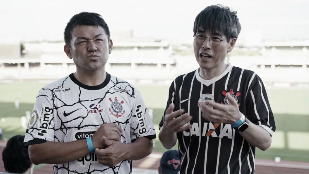 Vai Corinthians (2023) movie still: medium — Two men stand in a soccer stadium concourse overlooking the playing field. They are dressed in matching…