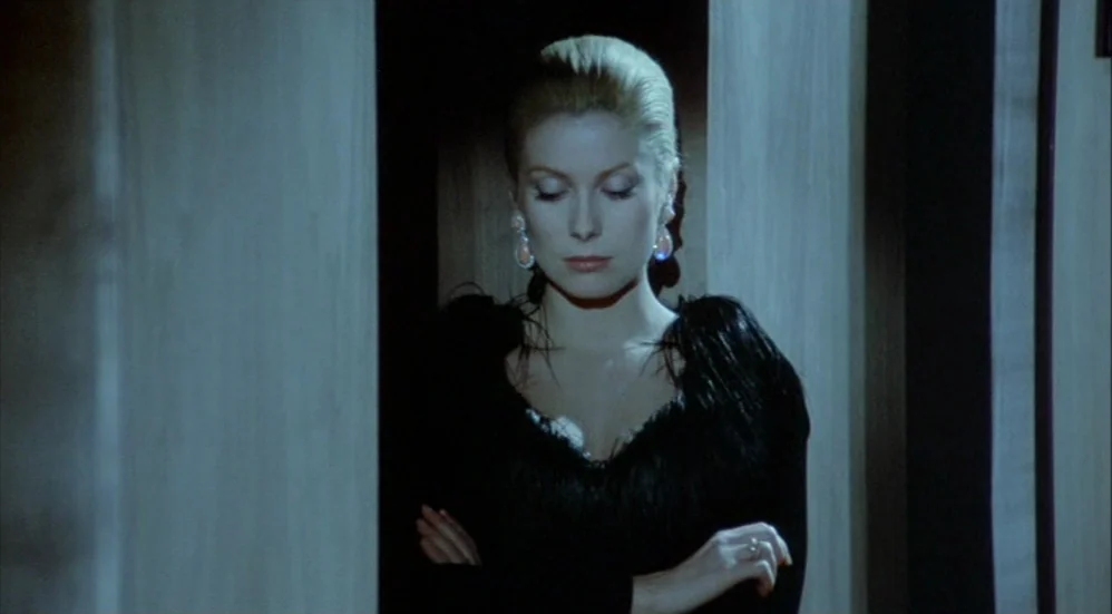Un Flic (1972) movie still: medium — A woman stands centered in a dark doorway framed by light-colored wooden panels. She wears a black feathered…